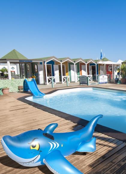 Pool mit Rutsche und haiförmigem Schwimmer, bunte Kabinen im Hintergrund.